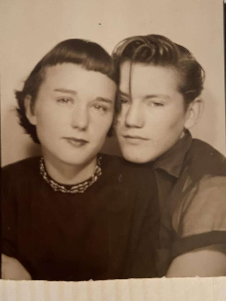 A black-and-white photograph shows a young woman with short hair and a necklace, and a young man with slicked-back hair. The woman is on the left and the man is on the right, closely leaning into each other, giving a sense of intimacy.