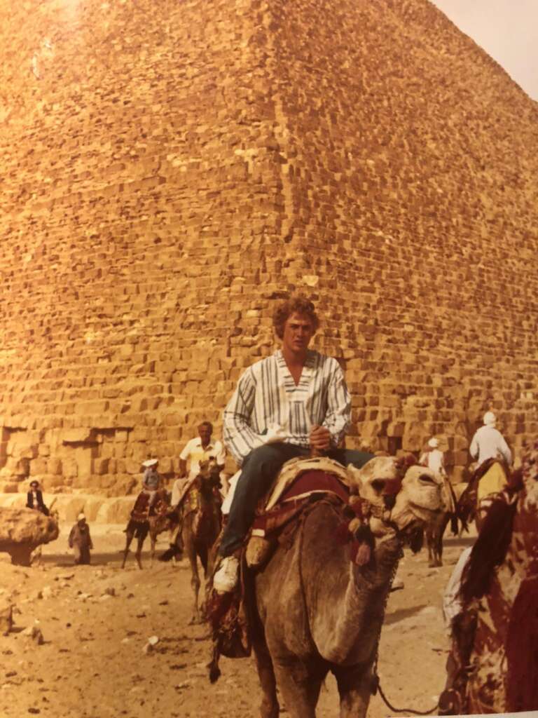 A person wearing a striped shirt is riding a camel in front of a large pyramid. Several other people and camels are visible in the background. The scene appears to be taken in a desert area.