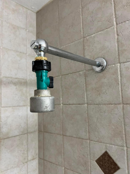 A showerhead pipe without a showerhead attached. The pipe is extended from the tiled wall, and there is a valve or adapter connected to the end of the pipe where the showerhead would typically be fixed. The wall tiles are creamy beige with a diamond-pattern accent.