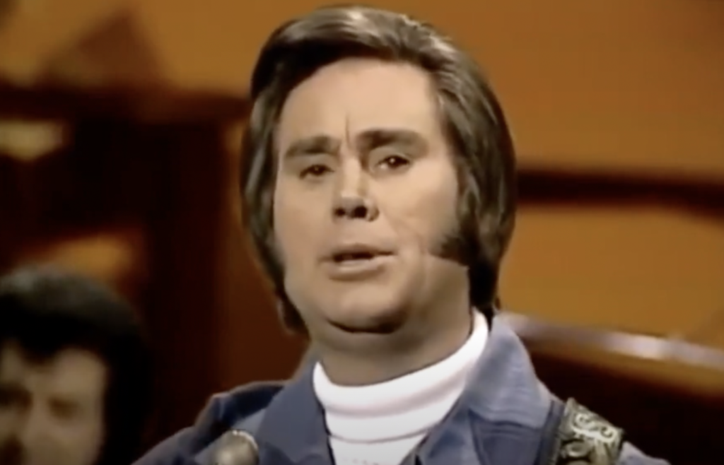 A man with neatly styled brown hair is singing into a microphone. He is wearing a white turtleneck underneath a blue blazer. The background is a warm, softly lit setting with blurred details.