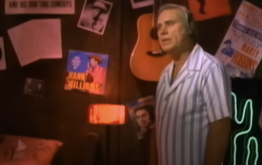 A man in striped pajamas stands in a dimly lit room with posters, a guitar on the wall, and various decorations. The ambiance suggests a retro or vintage theme with a spotlight highlighting the man.