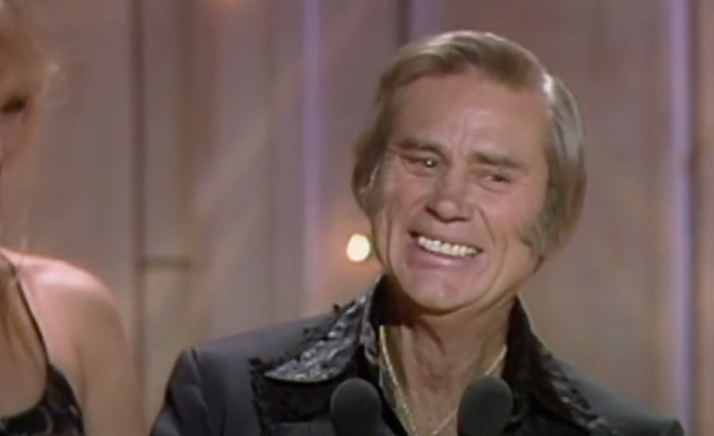 An older man with gray hair smiles broadly while standing at a microphone. He is dressed in a dark suit with a decorative collar. Another person stands partially visible beside him. The background features soft lighting and vertical panels.