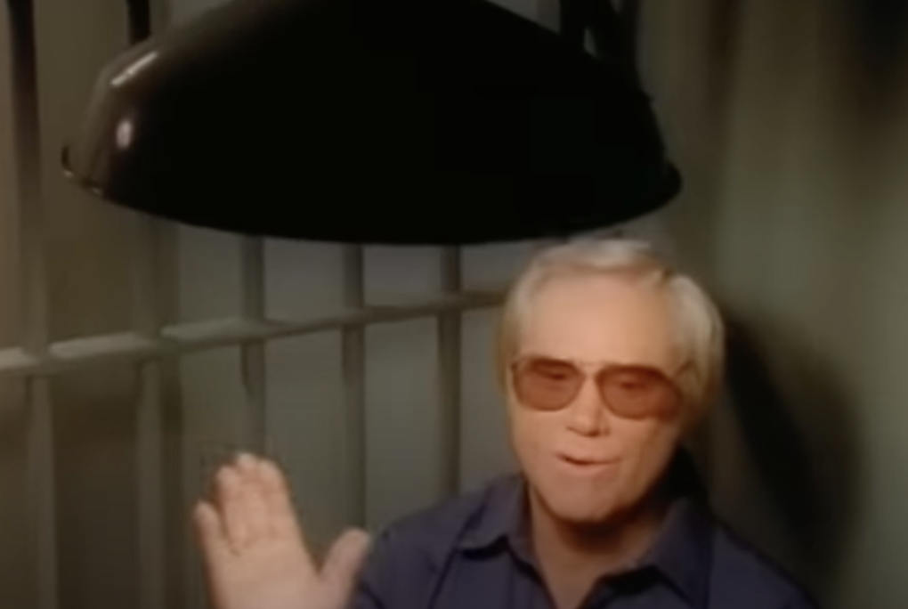 A man with grey hair and wearing sunglasses is waving while sitting under a large dark dome-shaped light. Behind him are vertical bars suggesting he is possibly in a jail cell or a similar setting.