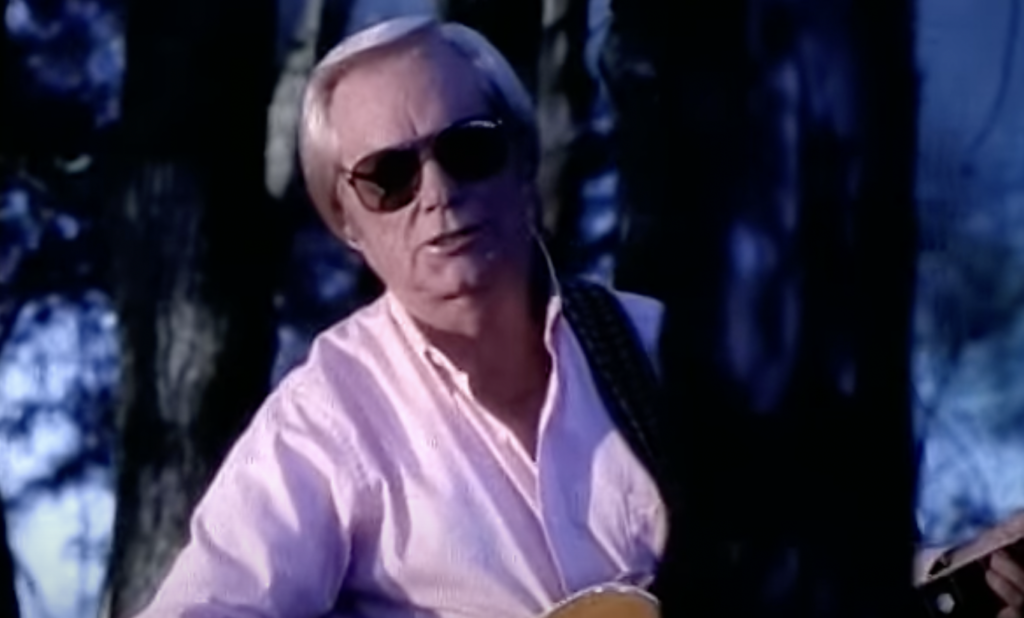 A man with short blonde hair and sunglasses is standing outdoors against a background of trees. He is wearing a light pink button-up shirt and holding a guitar close to his chest. The scene appears to be taken during twilight.