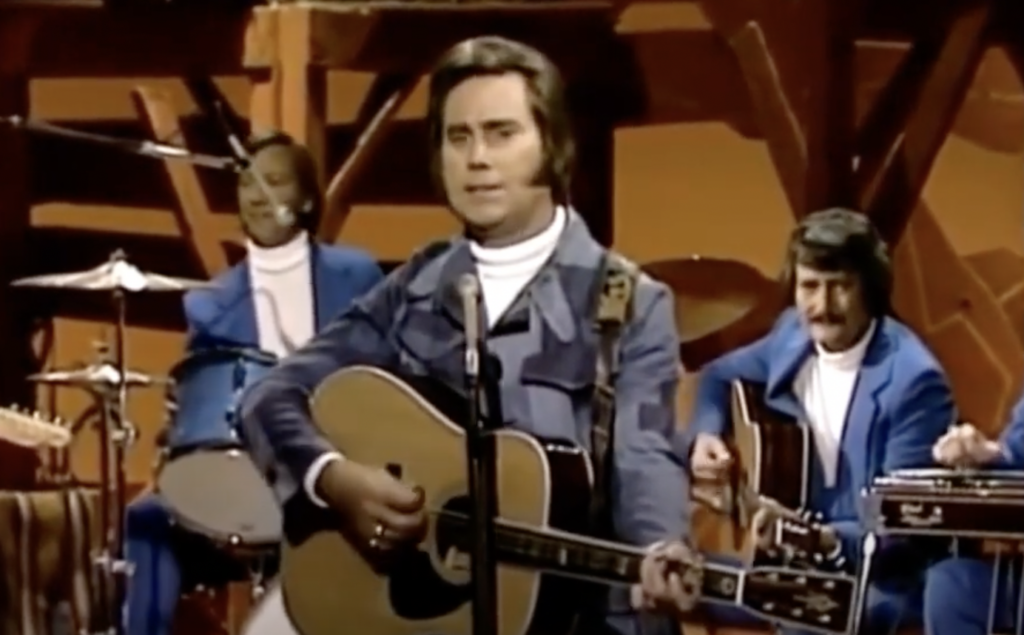 A male performer is playing an acoustic guitar and singing into a microphone on stage. He is accompanied by a drummer and another guitarist, all dressed in matching blue outfits with white turtlenecks. The backdrop is wooden with warm lighting.