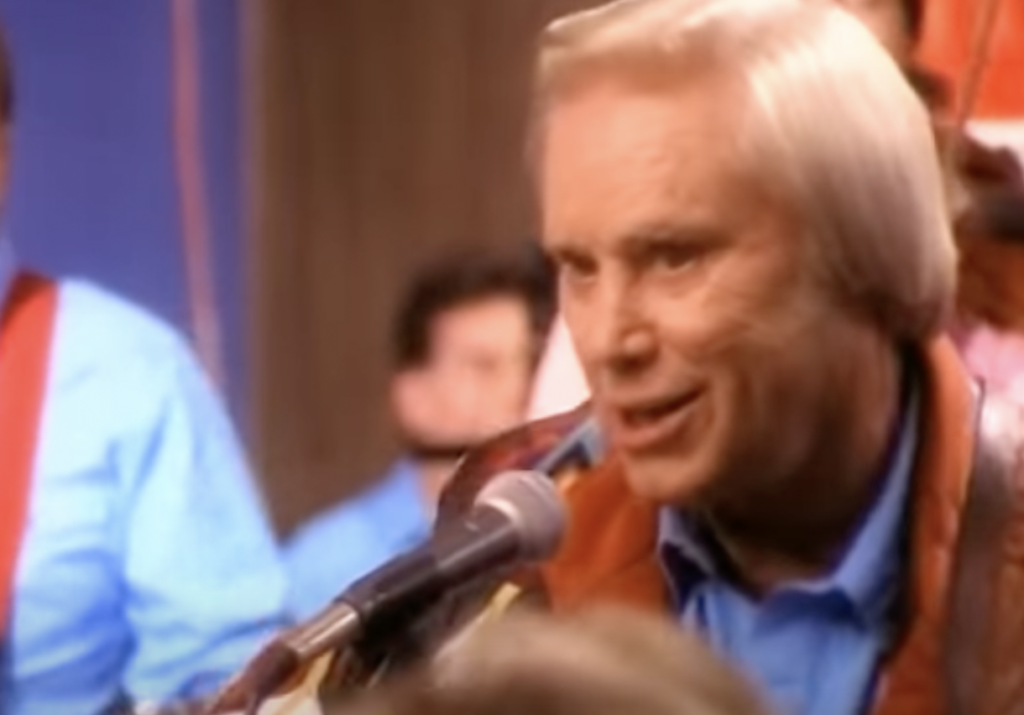 An older man with short, light-colored hair and a brown leather jacket sings into a microphone. The background features several out-of-focus individuals, some of whom are dressed in bright blue shirts.