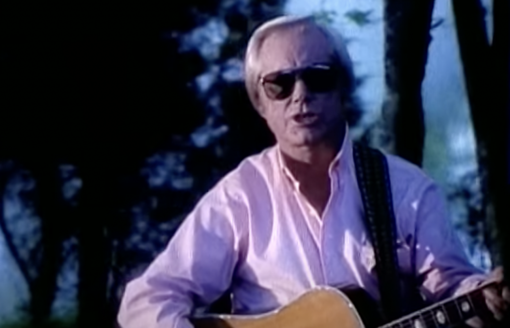 A person wearing sunglasses and a light pink dress shirt is sitting outdoors, holding an acoustic guitar. The background consists of trees and appears to be a forested area with dappled sunlight filtering through the leaves.