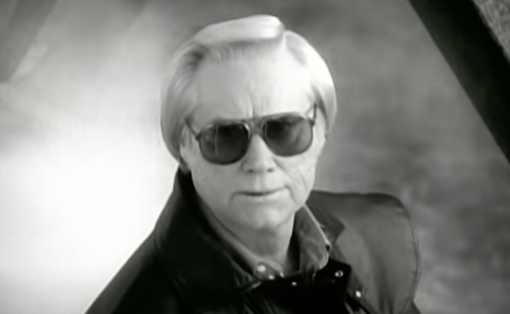 Black and white close-up of an older man with light-colored hair wearing sunglasses. He has a serious expression and is dressed in a leather jacket over a collared shirt. The background is a blurred, outdoors scene.