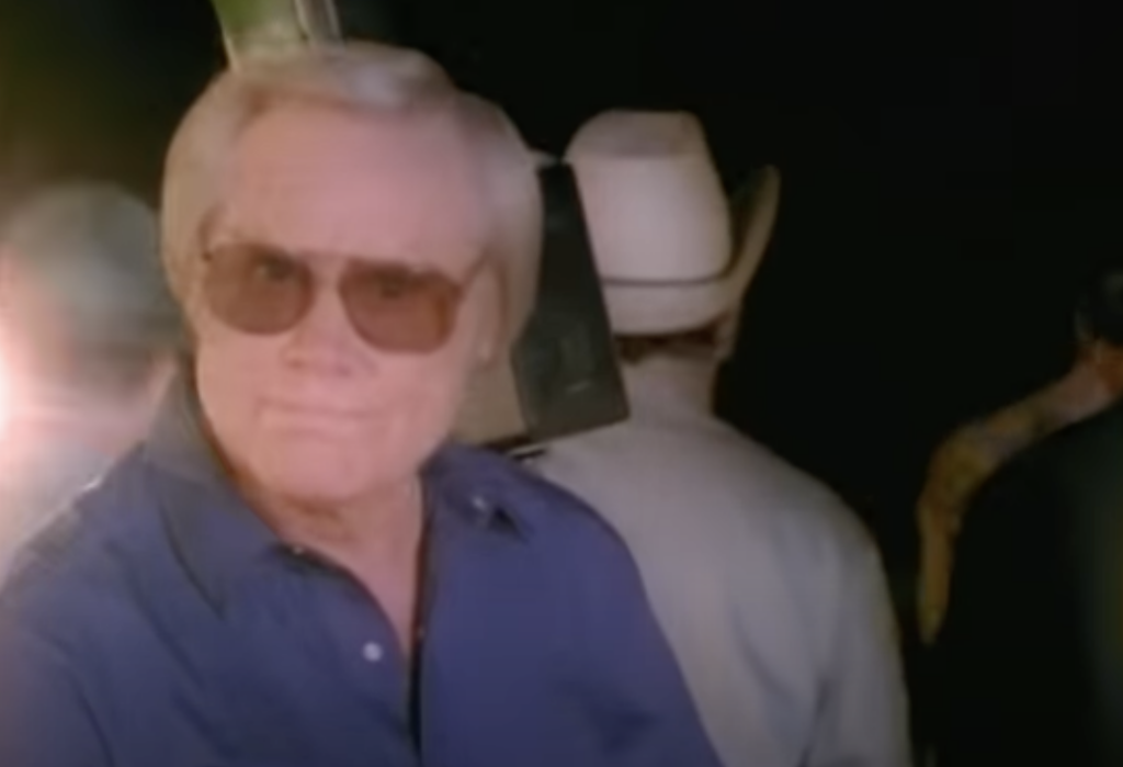 A man with light-colored hair, wearing sunglasses and a blue shirt, is looking at the camera. In the background, a person wearing a white cowboy hat is visible among a few other indistinct figures. The scene appears to have dim lighting.