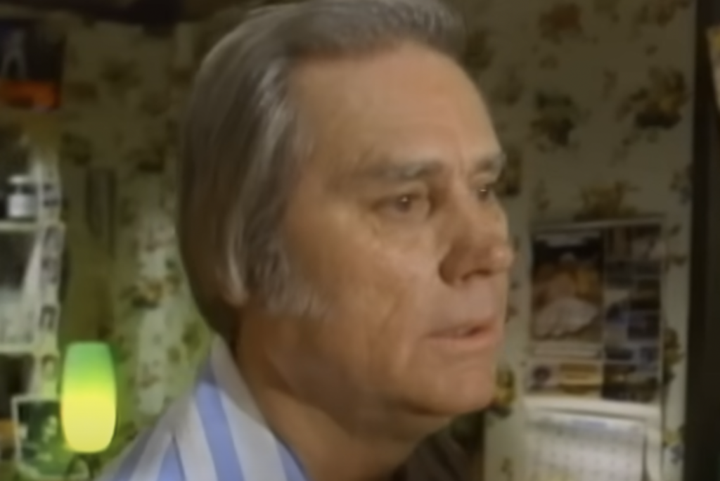 A contemplative older man with gray hair and wearing a blue and white striped shirt is standing indoors. Behind him is a wall with floral wallpaper, a green lamp, some shelves with books, and a calendar. The lighting suggests it is nighttime.