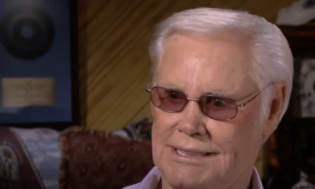 An elderly man with white hair and wearing tinted glasses is smiling. He is indoors, with a room featuring some framed items and decor in the background.