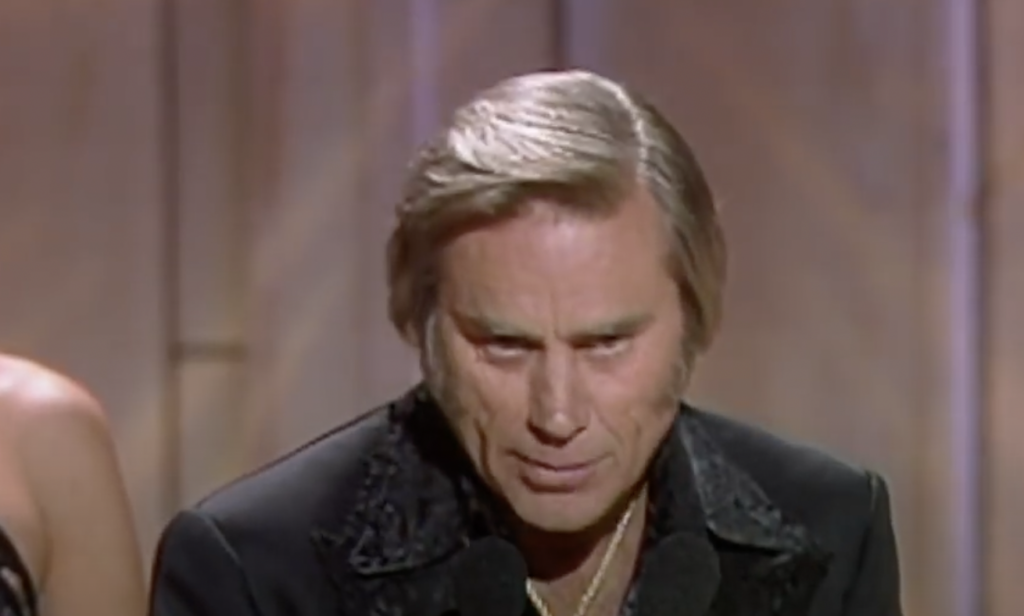 A man with short, slicked-back hair and wearing a black suit is seen standing and speaking into a microphone. The background appears to be a stage with a wooden panel backdrop. The lighting is warm, highlighting his facial features.