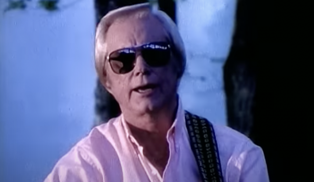 A person with light hair wearing dark sunglasses and a pink shirt is seen in close-up against a blurred outdoor background. They are playing a guitar, visible by the strap over their shoulder. The scene suggests a casual or relaxed setting.