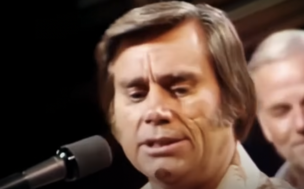 A man with short brown hair and sideburns is singing into a microphone. Another older man with white hair can be seen in the background. The scene appears to be indoors with the focus on the singer's face.