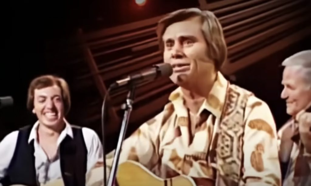 Three men are on stage performing. The man at the center is playing an acoustic guitar and singing into a microphone. He is wearing a patterned shirt. The man on the left is smiling, and the man on the right is playing a stringed instrument. The background is dimly lit.