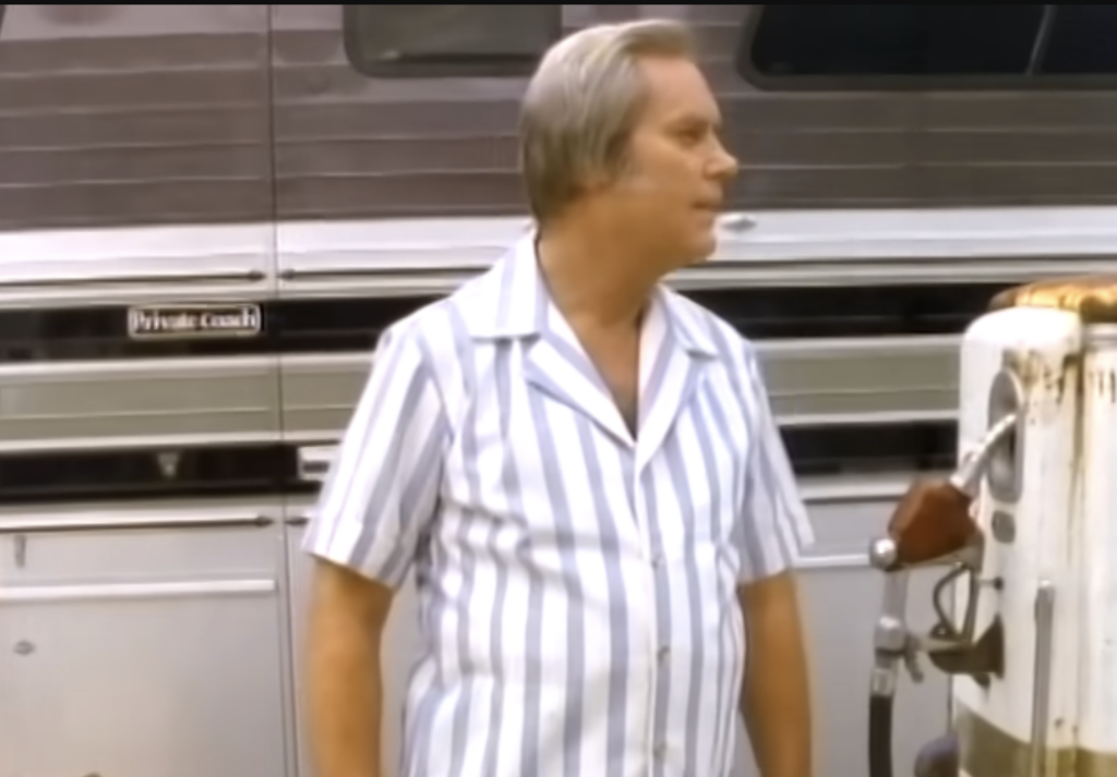 A person with graying hair stands beside an old-fashioned gas pump. They are wearing a light-blue and white vertically striped short-sleeve shirt. A large bus, labeled "Private Coach," is visible in the background.