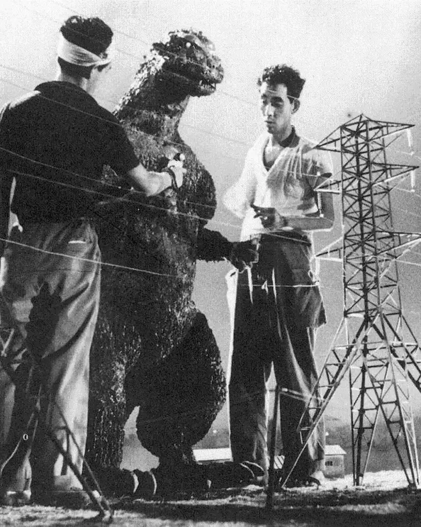 Black and white photo showing two men working on a life-sized Godzilla model on a film set. They are adjusting its body, standing close to a model of an electrical tower. Both appear focused, as they interact with the detailed and large monster figure.