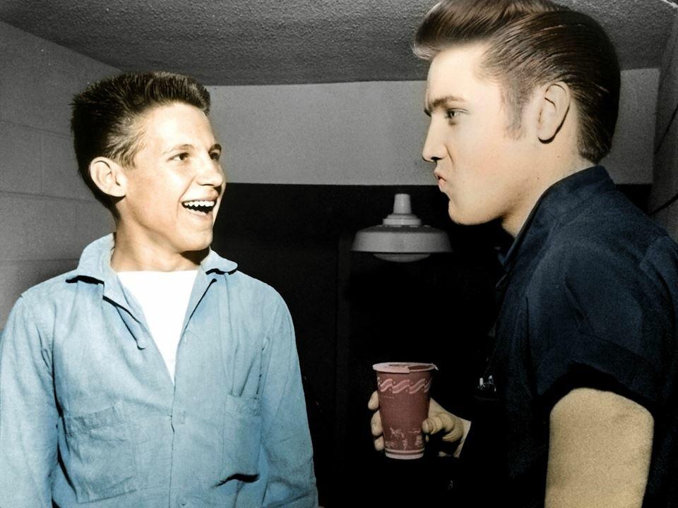 A young man with short hair, dressed in a light blue shirt, laughs and looks at another young man with styled hair, dressed in a dark blue shirt, who is holding a red paper cup and appears to be talking. The background is a plain indoor setting.