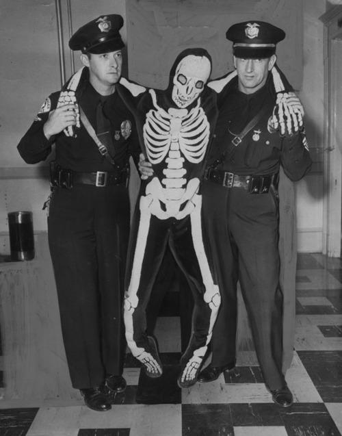 Two police officers in uniform stand on either side of a person wearing a skeleton costume, with their arms around the person. The scene appears to be indoors, with checkered flooring and a bench visible in the background.