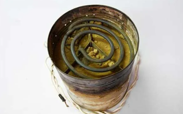 A cylindrical tin can with a rusty appearance is filled with a coiled heating element and wires protruding from it, resembling a makeshift or repurposed heater. The can's interior shows signs of corrosion and debris.