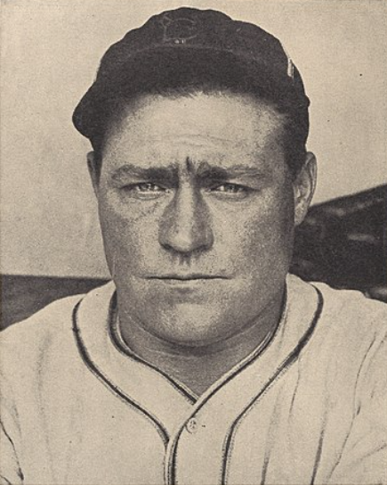 A black-and-white portrait of a man in a baseball uniform and cap, looking directly at the camera with a serious expression. The background is indistinct, suggesting it might be a sports venue or dugout.