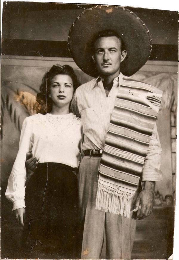 A black and white photo of a couple. The man is wearing a large sombrero and a traditional striped serape draped over one shoulder. The woman, standing beside him with her arm around his back, is dressed in a white blouse and dark skirt. Both look at the camera.