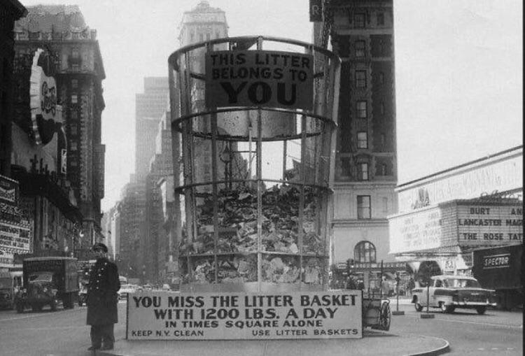 A black and white photo of Times Square in the mid-20th century featuring a towering, transparent litter basket filled with trash. A sign on the basket reads, "This litter belongs to YOU". Below, another sign states, "You miss the litter basket with 1200 lbs. a day in Times Square alone". A policeman stands near the basket and cars are visible in the background.