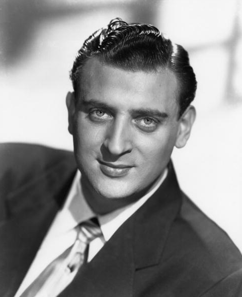 Black and white portrait of a man with neatly combed, dark hair and light eyes, wearing a suit and tie. He faces the camera with a slight smile and confident expression. The background is blurred.