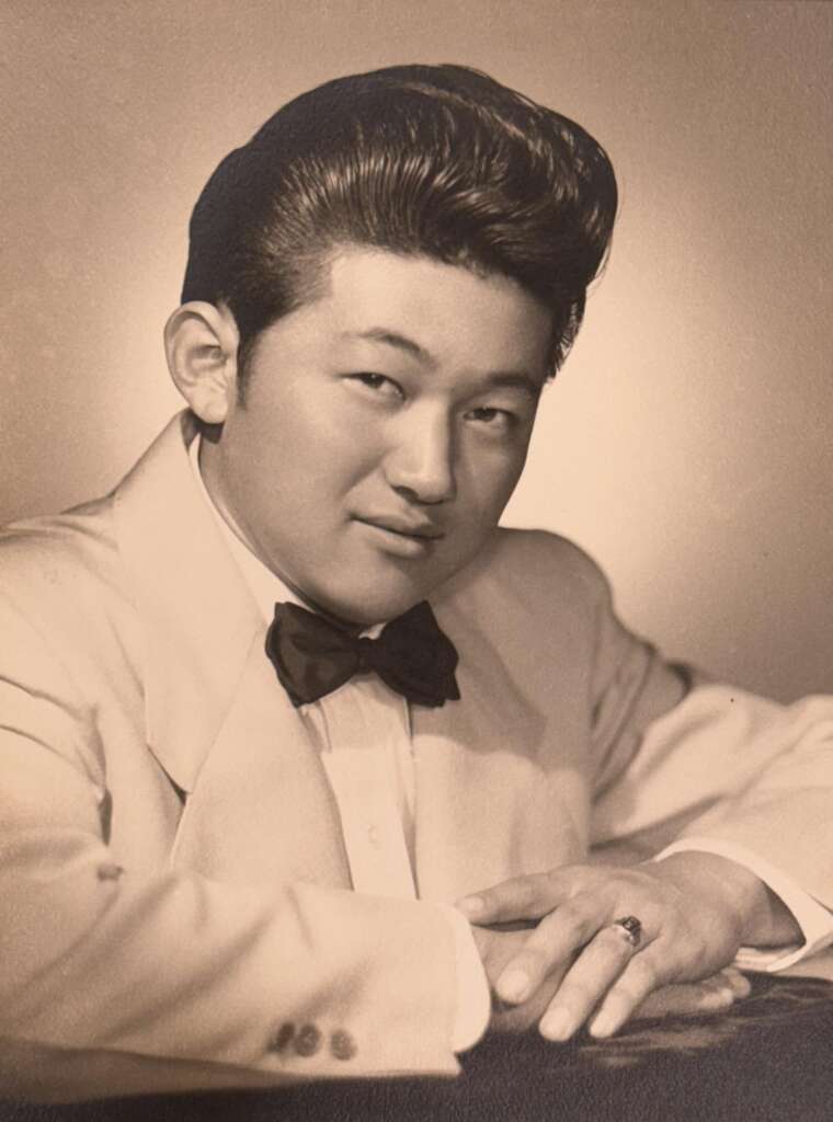 A vintage black-and-white portrait of a young man wearing a tuxedo with a bow tie. He has neatly styled hair and is resting his arms on a surface while looking slightly to the side with a gentle smile.