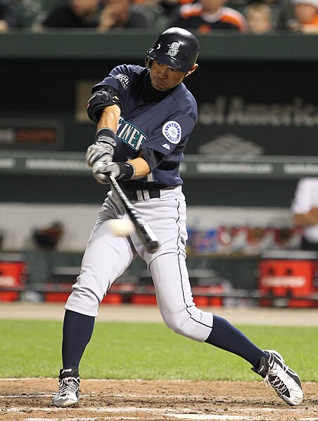 A baseball player in a navy blue and gray uniform swings a bat during a game, making contact with the ball. The player is in mid-action with a focused expression, and the dugout and spectators are visible in the background.