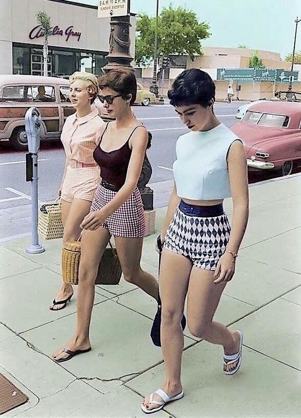 Three women walk down a street in vintage summer outfits, including shorts and sleeveless tops. They pass by parked cars and a parking meter. One woman carries a woven bag. The scene appears to be from the mid-20th century, with retro cars and building signage.