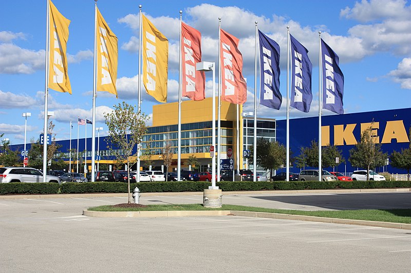 A large IKEA store with a blue and yellow facade is seen on a sunny day. Numerous yellow and blue IKEA flags are suspended on flagpoles in front of the building. Cars are parked in the parking lot, and a clear blue sky with scattered clouds is in the background.