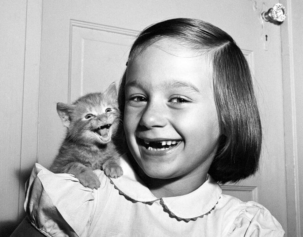 A young girl with a bob haircut, missing some front teeth, smiles widely while holding a tiny, yawning kitten on her shoulder. Both appear cheerful and full of joy against a plain background.