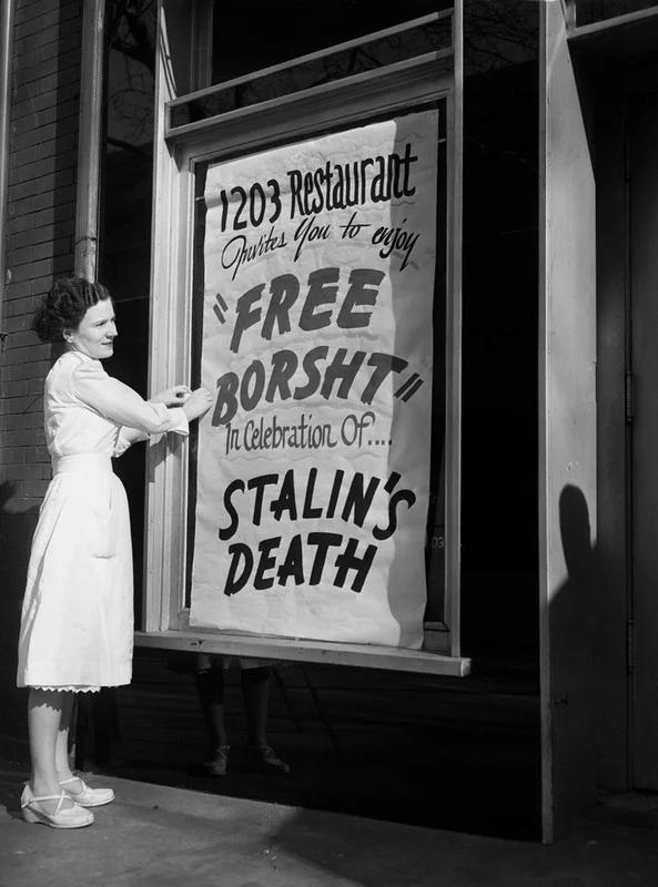 A woman in a white dress stands by a large window, holding a sign that reads, "1203 Restaurant invites you to enjoy ‘Free Borsht’ in celebration of... STALIN'S DEATH." The sign is displayed in the window of the restaurant, and the woman appears to be smiling.