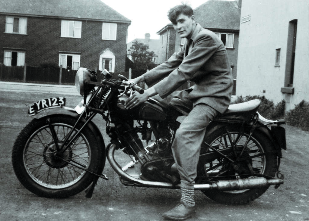 A person wearing a jacket and pants is sitting on a vintage motorcycle with the license plate EYR 123. The background shows houses with brick walls and fenced yards on a quiet street. The image has a black and white, nostalgic appearance.