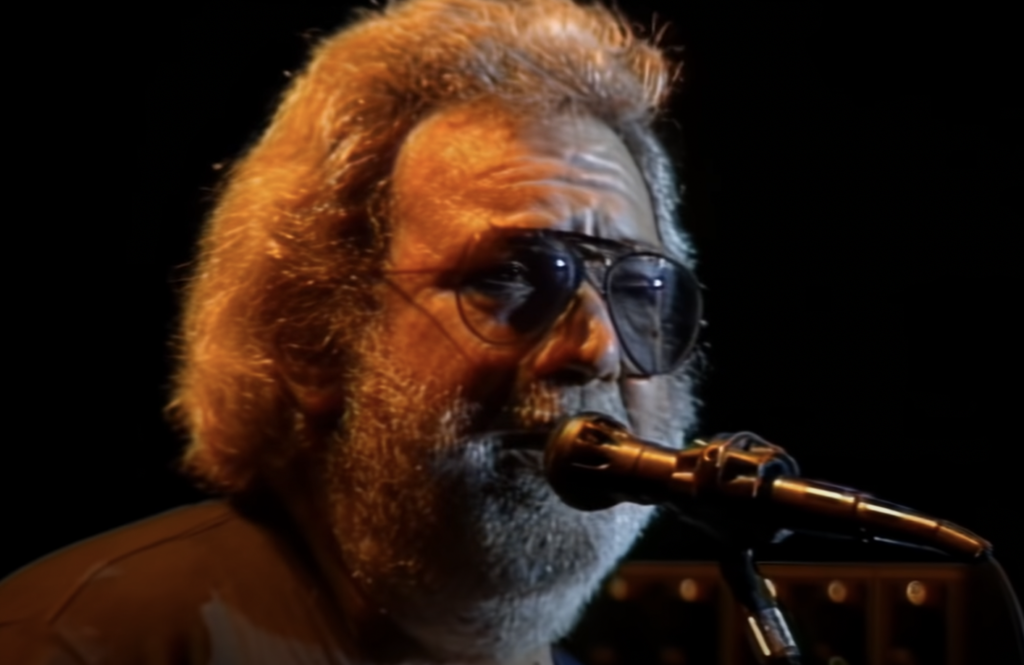 A man with a grey beard and long, slightly tousled hair, wearing dark sunglasses, sings into a microphone. The background is dark, emphasizing his face and the microphone stand in front of him.