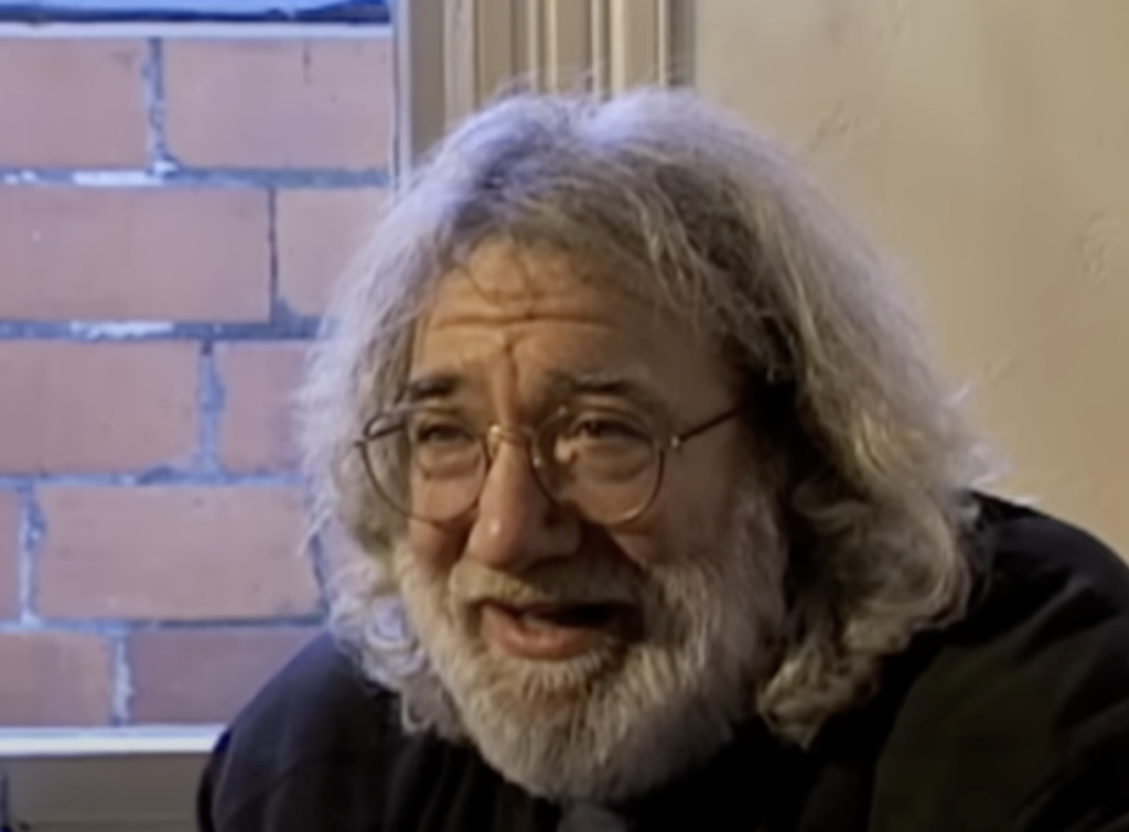 A man with long gray hair, a beard, and glasses is smiling. He is sitting indoors near a window with a red brick wall visible outside. The lighting is soft, creating a warm and relaxed atmosphere. He appears to be in mid-conversation.