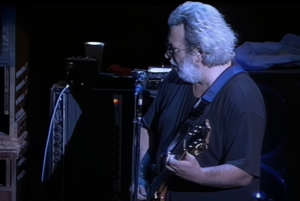 A man with white hair and glasses is playing an electric guitar on stage. He's wearing a black shirt and is focused on his performance with a microphone stand in front of him. The stage background is dark, creating a contrast with the subject.