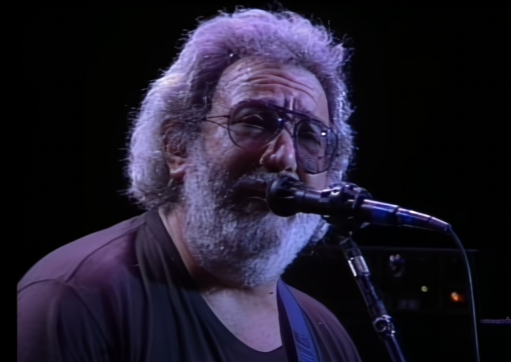 A man with gray hair and a beard, wearing eyeglasses and a dark shirt, sings into a microphone on a dimly lit stage. The background is mostly dark, emphasizing the focus on the singer.