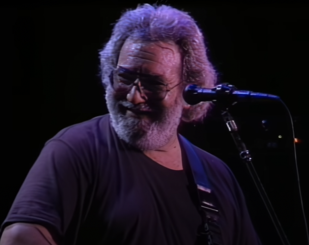 A bearded man with glasses and long hair smiles while playing an electric guitar. He wears a dark T-shirt and is illuminated by stage lights, with a microphone positioned in front of him, suggesting he is performing music.