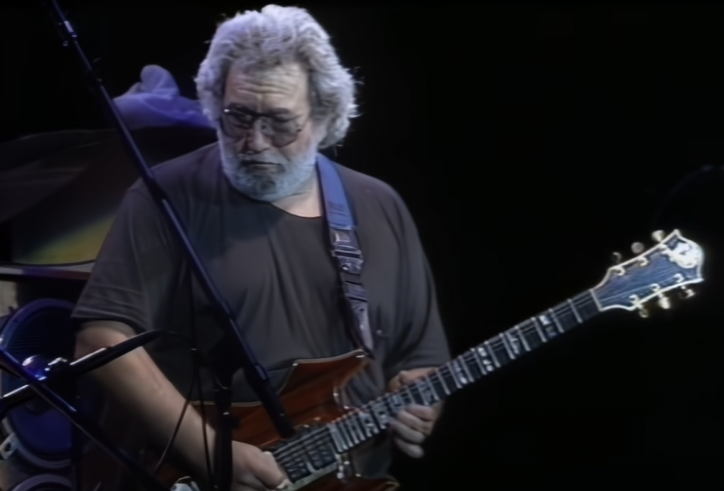 A man with grey hair and a beard is playing an electric guitar on stage. He is wearing sunglasses and a dark T-shirt. Musical equipment and a microphone stand are visible in the background. The scene is lit with stage lighting.