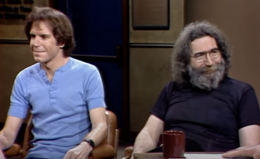 Two men are seated together in a room. The man on the left is wearing a light blue shirt, while the man on the right has a bushy beard, long hair, and wears a black shirt. They appear to be engaged in a conversation, with a red mug on the table in front of them.