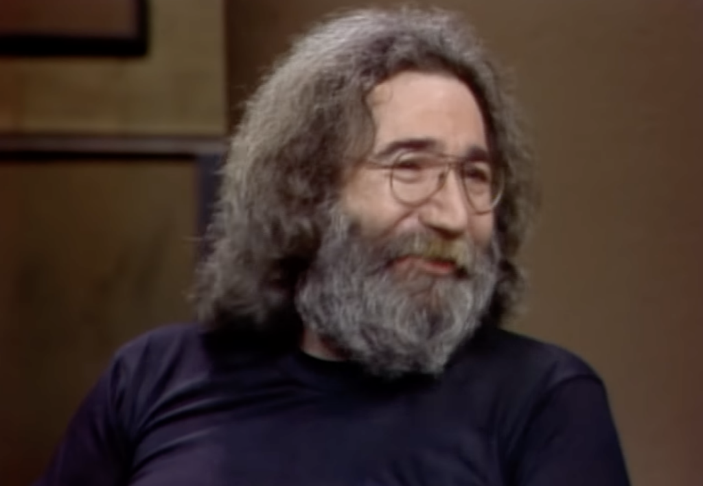 A man with long, gray hair and a full beard, wearing glasses and a black shirt, is shown smiling in an indoor setting. The background appears out of focus.