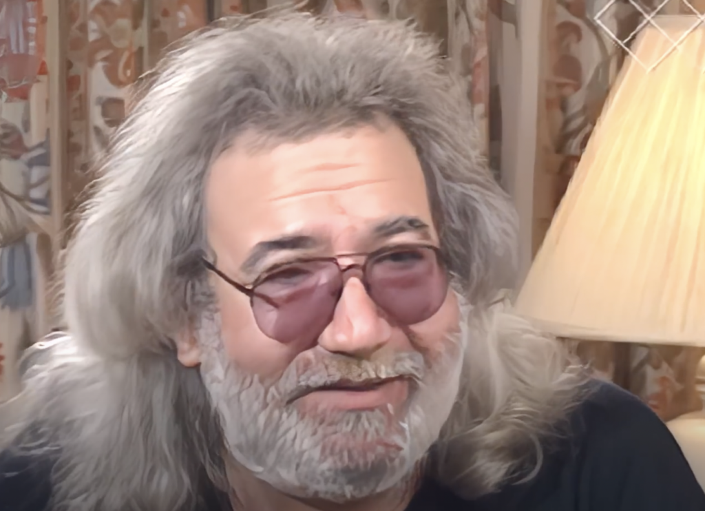 A person with long gray hair, a gray beard, and wearing round purple-tinted glasses is smiling. There is a lamp with a beige shade and a floral curtain in the background.