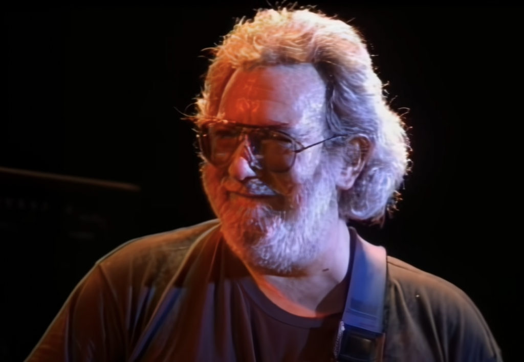 A man with gray, wavy hair, a thick beard, and glasses is smiling and looking off to the side. He is wearing a dark-colored T-shirt and has a guitar strap over his shoulder. The background is dark, accentuating the lighting on his face and hair.