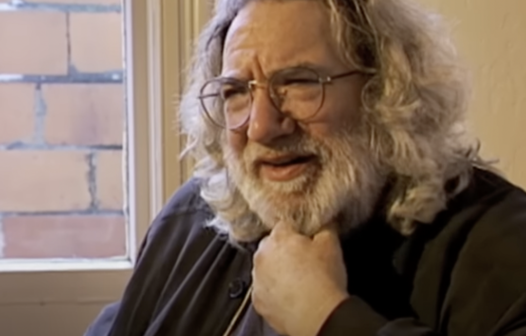 A person with long, curly gray hair, glasses, and a thick beard is seen indoors. They are resting their chin on their hand and looking slightly to the side. A window with a view of a brick wall is in the background.