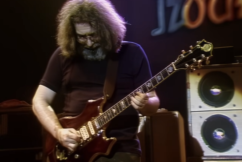 A musician with long, wavy hair and glasses is playing an electric guitar on stage. The background is dimly lit with a blurry sign and large speaker cabinets. The musician appears deeply focused on their performance.