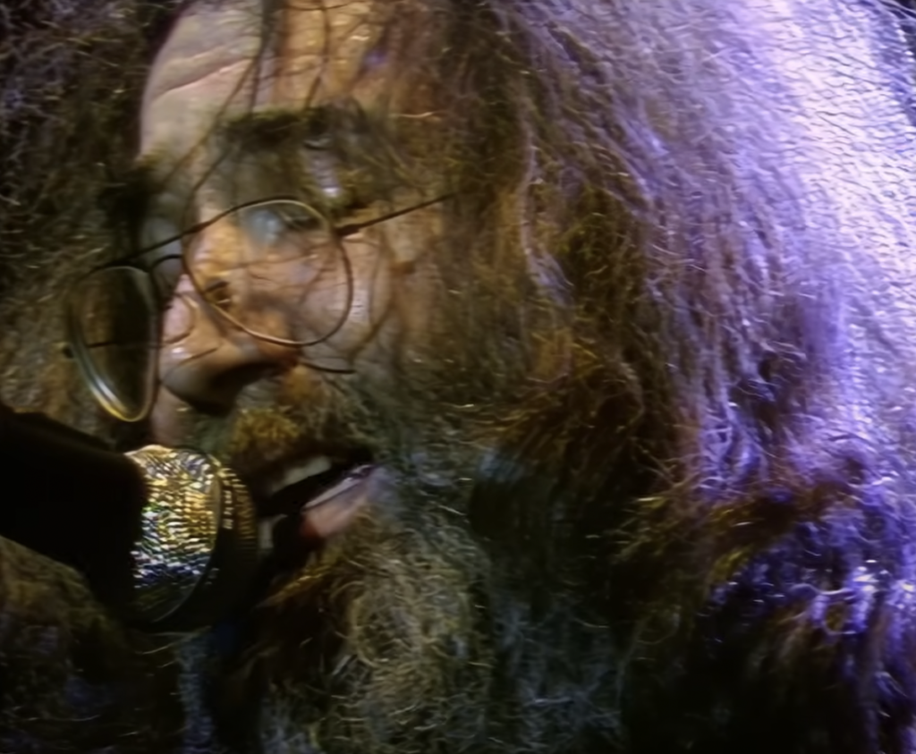 A close-up of a person with long, thick, curly hair and a full beard, wearing round glasses, singing into a microphone under purple stage lights. The person's face shows intense emotion as they perform.