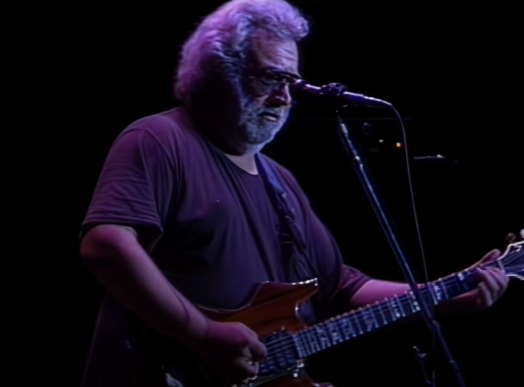 A person with gray hair, beard, and glasses is playing an electric guitar and singing into a microphone on stage against a black backdrop. The person is wearing a dark, short-sleeve shirt and appears to be deeply focused on their performance.