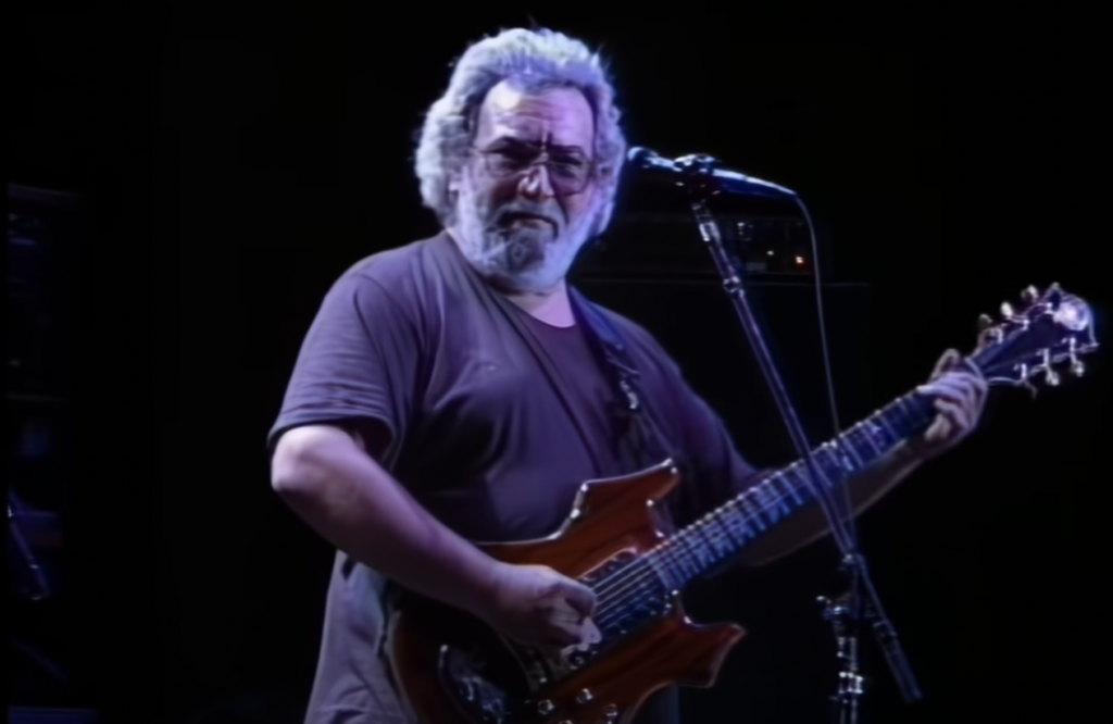 A man with gray hair and glasses is playing an electric guitar on stage. He is standing in front of a microphone and is wearing a dark-colored t-shirt. The background is dimly lit, emphasizing the performer.